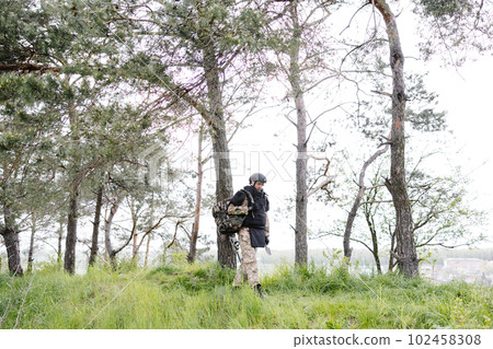 Young soldier in uniforms and tactical vest works in the forest and prepares for action at a temporary forest base. A man does in the work of demining the territory Young soldier in uniforms and tactical vest works in the forest and prepares for action at a temporary forest base. A man does in the work of demining the territory 102458308