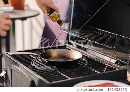 Close-up of man add olive oil on frying carbon steel pan which is on the grill. Close-up of man add olive oil on frying carbon steel pan which is on the grill. 102458353