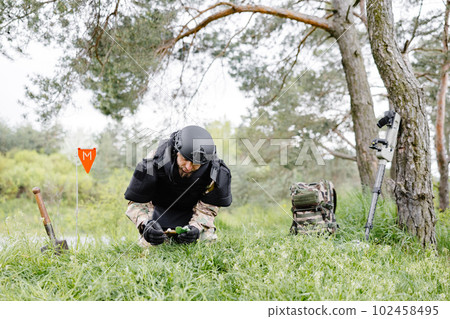 A man in a military uniform and a bulletproof vest works in the forest to demine the territory. A man warns of danger by making a red mark A man in a military uniform and a bulletproof vest works in the forest to demine the territory. A man warns of danger by making a red mark 102458495