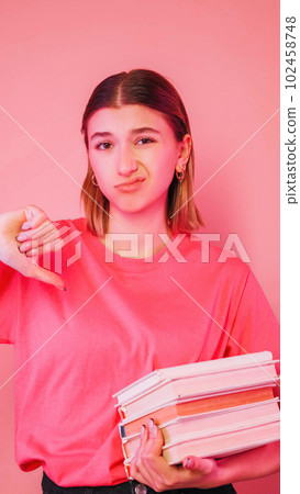 Dislike student. Boring education. Exam failure. Dissatisfied sad girl holding stack of books showing thumb down sign grimacing isolated on neon color pink background. 102458748