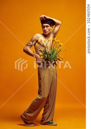 Full-length portrait of stylish, handsome, young man posing shirtless in beret and beige pants against yellow studio background Full-length portrait of stylish, handsome, young man posing shirtless in beret and beige pants against yellow studio background 102460504