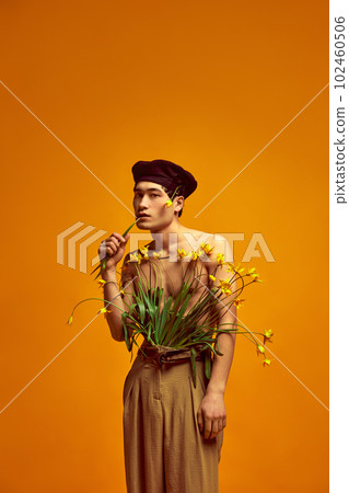Comfort and style. Portrait of young handsome man in black beret, classical beige pants, posing shirtless with flowers against yellow studio background Comfort and style. Portrait of young handsome man in black beret, classical beige pants, posing shirtless with flowers against yellow studio background 102460506