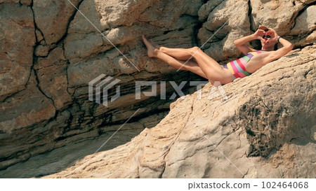 A beautiful slim woman in striped swimsuit makes heart shape with her hands while sunbathing on rocks at the beach 102464068