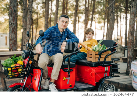 Farmer father and school boy kid delivered wooden crates with vegetables to camp site for picnic. Dad man male and son tourists delivered groceries on electric tricycle vehicle in the forrest. Farmer father and school boy kid delivered wooden crates with vegetables to camp site for picnic. Dad man male and son tourists delivered groceries on electric tricycle vehicle in the forrest. 102464477