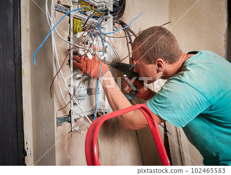 Side view of man in work gloves fixing electrical switchboard with electric drill in apartment under renovation. Electrician repairing household electrical panel with wires and switches. Side view of man in work gloves fixing electrical switchboard with electric drill in apartment under renovation. Electrician repairing household electrical panel with wires and switches. 102465583