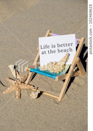 LIFE IS BETTER AT THE BEACH text on paper greeting card on background of beach chair lounge starfish summer vacation decor. Sandy beach sun. Holiday concept postcard. Travel LIFE IS BETTER AT THE BEACH text on paper greeting card on background of beach chair lounge starfish summer vacation decor. Sandy beach sun. Holiday concept postcard. Travel 102469628