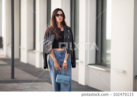 Portrait of gorgeous brunette woman standing city street. Fashion model wears black leather jacket, jeans, short top, small handbag on chain. Street style Portrait of gorgeous brunette woman standing city street. Fashion model wears black leather jacket, jeans, short top, small handbag on chain. Street style 102470115