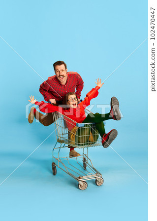 Portrait with couple, man and pretty woman having a shopping cart race over blue color studio background. Adventure time Portrait with couple, man and pretty woman having a shopping cart race over blue color studio background. Adventure time 102470447