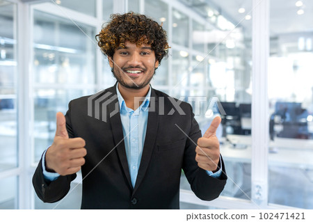 Portrait of successful hispanic businessman boss, manager in business suit and beard looking at camera and smiling standing near window, showing thumbs up, sign of success and achieving goals. Portrait of successful hispanic businessman boss, manager in business suit and beard looking at camera and smiling standing near window, showing thumbs up, sign of success and achieving goals. 102471421
