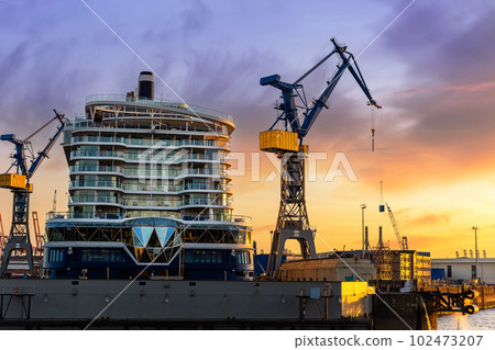 Scenic back view large modern luxury cruise ship liner under construction building cranes at dry dock shipyard in Hamburg port dramatic sunset sky background. Big vessel manufactoring site industry 102473207