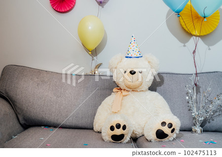 Big Teddy bear with balloons in party cap sitting on the gray sofa in decorated room. Birthday or festive holiday background. Selective focus, copy space Big Teddy bear with balloons in party cap sitting on the gray sofa in decorated room. Birthday or festive holiday background. Selective focus, copy space 102475113
