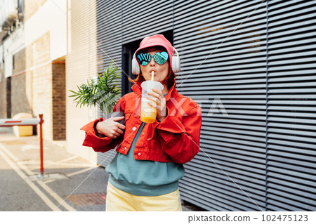 Hipster fashion woman in bright clothes, heart shaped glasses, headphones, bucket hat drinking fruity flavored tapioca bubble tea and holding green potted plant on the gray striped wall background Hipster fashion woman in bright clothes, heart shaped glasses, headphones, bucket hat drinking fruity flavored tapioca bubble tea and holding green potted plant on the gray striped wall background 102475123