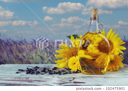A bottle of oil, fresh sunflower flowers and seeds on the table, against the backdrop of a landscape with a flowering field, organic products, harvest, farm, sale, agriculture A bottle of oil, fresh sunflower flowers and seeds on the table, against the backdrop of a landscape with a flowering field, organic products, harvest, farm, sale, agriculture 102475901