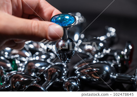 A woman holds a metal anal plug with a blue crystal against the background of many metal anal plugs on a black silk sheet.  102478645