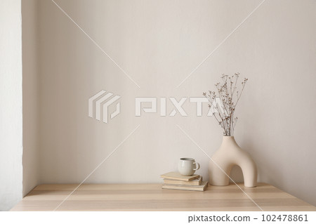 Elegant interior still life. Modern organic, geometric shaped vase with dry flowers, grass. Pile of old books, cup of coffee on wooden table. Home staging, minimal decor concept. Blank beige wall. 102478861