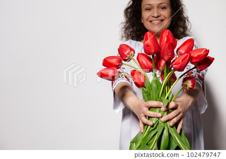 Details on a bouquet of red tulips in the hands of a beautiful smiling woman, dressed in Ukrainian traditional embroidered dress, isolated on white color background. Ukrainian culture and folklore 102479747