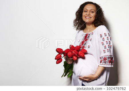Delightful adult pregnant woman, expectant mother dressed in Ukrainian ethnic embroidered clothing, holding her belly, posing with a bouquet of red tulips, isolated on white background. Copy ad space Delightful adult pregnant woman, expectant mother dressed in Ukrainian ethnic embroidered clothing, holding her belly, posing with a bouquet of red tulips, isolated on white background. Copy ad space 102479762