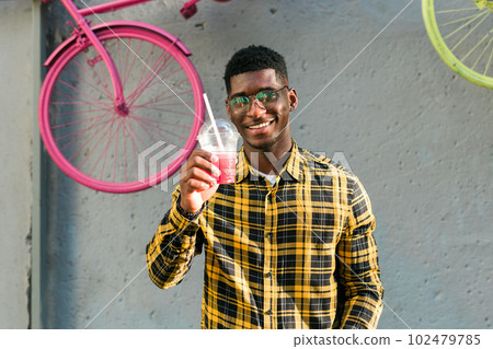 Carefree african american guy in casual wear holding tropical cocktail urban summer background. Cool young man drinking yummy alcohol-free beverage on summer vacation 102479785