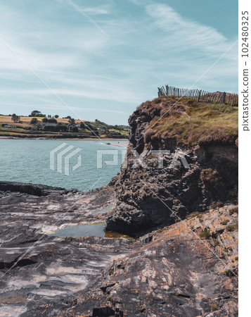 A sheer stone cliff on the Atlantic Ocean in Ireland on a sunny summer day. Irish seaside landscape, clear blue sky, rock formation near water. 102480325