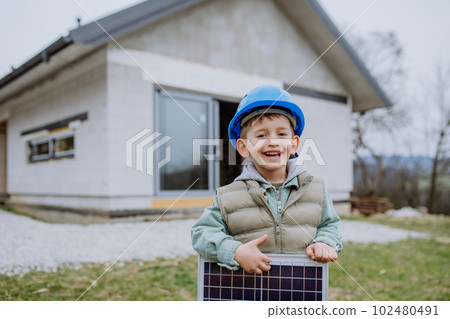 Portrait of little boy holding solar panel, in front of their new unfinished house. Portrait of little boy holding solar panel, in front of their new unfinished house. 102480491