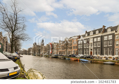 some boats in the water and buildings on the other side of the canal with cars parked along the river bank 102481783