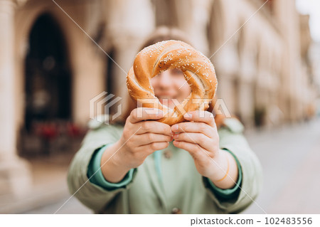 Attractive young female tourist is holding prezel, traditional polish snack on the Market square in Krakow. Traveling Europe in spring. Selective focus. High quality photo Attractive young female tourist is holding prezel, traditional polish snack on the Market square in Krakow. Traveling Europe in spring. Selective focus. High quality photo 102483556