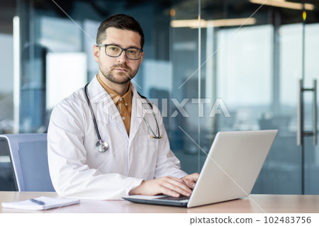 Portrait of a young serious doctor inside the office at the workplace of the clinic, the man is looking thoughtfully at the camera, the doctor is working with a laptop. Portrait of a young serious doctor inside the office at the workplace of the clinic, the man is looking thoughtfully at the camera, the doctor is working with a laptop. 102483756