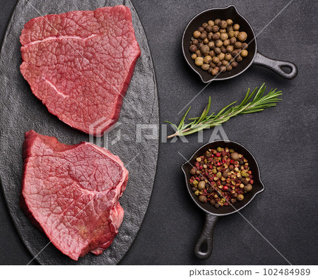 Raw piece of beef with spices pepper, rosemary sprig, salt and olive oil on a wooden board, black background Raw piece of beef with spices pepper, rosemary sprig, salt and olive oil on a wooden board, black background 102484989