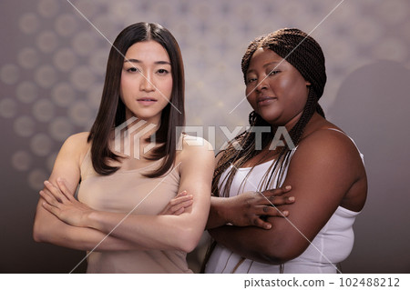 Confident diverse women posing with crossed hands, showing success gesture and looking at camera. African american and asian models with different body types standing with folded arms Confident diverse women posing with crossed hands, showing success gesture and looking at camera. African american and asian models with different body types standing with folded arms 102488212