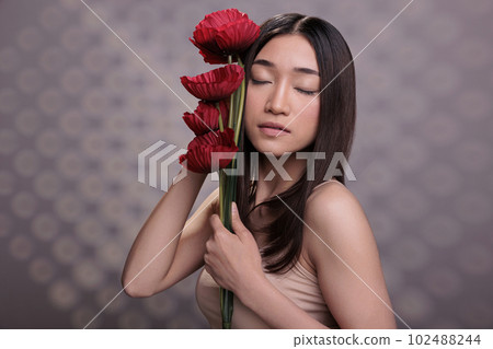 Asian woman with closed eyes holding red poppies branch. Beautiful young model wearing beige casual top, standing with summer flowers bouquet, showing skincare and body care concept 102488244