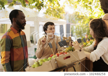 Young customers tasting free food samples at farmers market, fresh juicy fruits and vegetables outside. Man and woman buying healthy products from local fresh organic marketplace. Young customers tasting free food samples at farmers market, fresh juicy fruits and vegetables outside. Man and woman buying healthy products from local fresh organic marketplace. 102488258