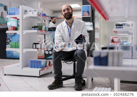 Smiling pharmacist holding medical tonometer ready to measure customers hypertension during checkup visit in pharmacy. Drugstore filled with supplements and drugs, medicine support service Smiling pharmacist holding medical tonometer ready to measure customers hypertension during checkup visit in pharmacy. Drugstore filled with supplements and drugs, medicine support service 102488276