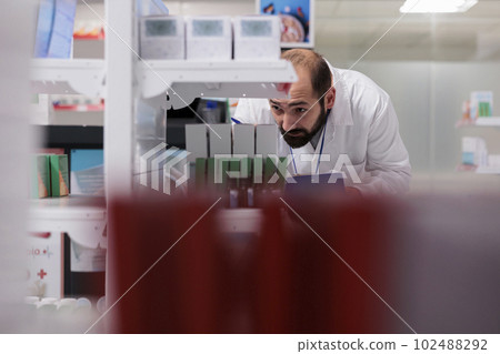 Pharmacist looking at shelves full with drugs packages doing inventory writing vitamins name on papers, working in drugstore. Pharmacy employee is responsible for reviewing orders to ensure accuracy. Pharmacist looking at shelves full with drugs packages doing inventory writing vitamins name on papers, working in drugstore. Pharmacy employee is responsible for reviewing orders to ensure accuracy. 102488292