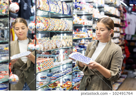 Curious female tourist looking to buy ornamental ceramic plates at souvenir shop Curious female tourist looking to buy ornamental ceramic plates at souvenir shop 102491764