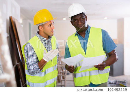 Foreman and worker in yellow vest and protective helmet are discussing the drawings of the repaired premises in cottage 102492239