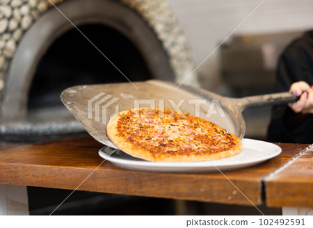 Female chef in black uniform prepares pizza and pulls it out of oven in kitchen of pizzeria 102492591