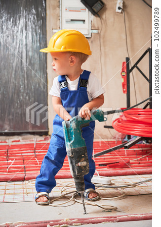 Child drilling floor with electric hammer drill at home during renovation. Kid construction worker using drilling machine while installing underfloor heating system in apartment. Child drilling floor with electric hammer drill at home during renovation. Kid construction worker using drilling machine while installing underfloor heating system in apartment. 102499789