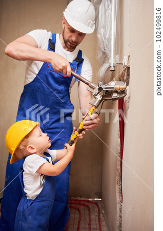 Man and child using pipe pressing tool while installing plumbing pipes in bathroom. Male plumber and kid in safety helmets working on sanitary equipment installation in apartment under renovation. 102499816