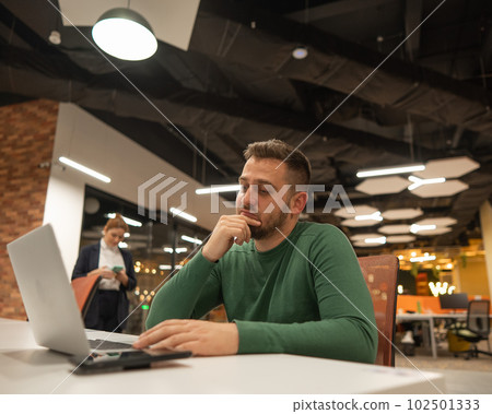 Bearded man holding his head while working on a laptop in an open space office. Red-haired caucasian woman uses smartphone.  102501333