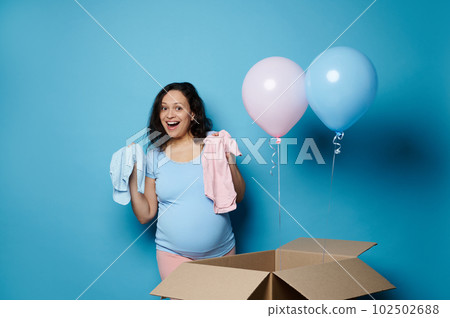 Happy pregnant woman with big belly, in 2nd semester of her pregnancy, showing a blue and pink newborn bodysuit, expressing amazement and gender reveal party, baby shower, isolated on blue background 102502688
