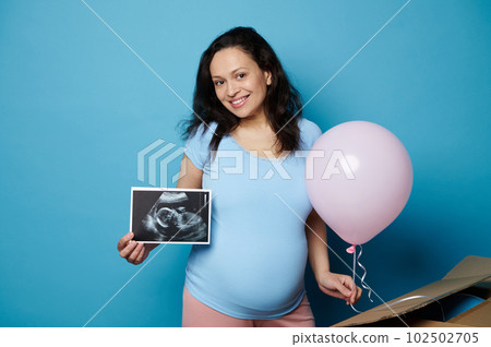 Portrait of dark haired multi ethnic cheery adult pregnant woman, showing an ultrasound scan of her newborn baby, smiling broadly looking at camera, holding pink balloon, isolated on blue background. Portrait of dark haired multi ethnic cheery adult pregnant woman, showing an ultrasound scan of her newborn baby, smiling broadly looking at camera, holding pink balloon, isolated on blue background. 102502705