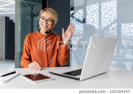 Portrait of successful tech support worker, woman with blonde hair smiling and looking at camera, businesswoman with headset for video call, greeting gesture, inside office with laptop. Portrait of successful tech support worker, woman with blonde hair smiling and looking at camera, businesswoman with headset for video call, greeting gesture, inside office with laptop. 102504147
