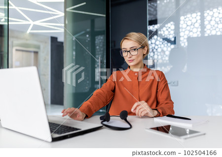 Serious and concentrated woman with video call headset looking at camera, blonde woman thinking close up inside office, call center and support tech worker . Serious and concentrated woman with video call headset looking at camera, blonde woman thinking close up inside office, call center and support tech worker . 102504166