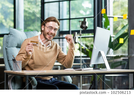 A young man successfully uses a credit card. He sits happily in the office at the computer, holds a card, looks at the camera, shows a yes gesture, celebrates. 102506213
