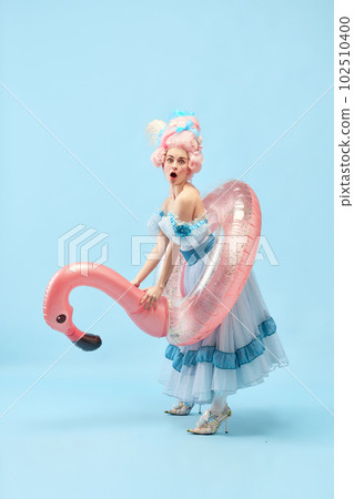 Portrait with funny beautiful princess, queen wearing dress holding pink inflatable flamingo and going beach over blue background 102510400