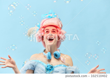 Portrait with princess, queen wearing dress playing with soap bubbles and smiling over blue studio background. Cheerful young girl 102510402