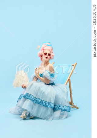 Portrait of charming princess, queen wearing fancy dress sitting on sunbed and using hand fan over blue background. Hot summer weather Portrait of charming princess, queen wearing fancy dress sitting on sunbed and using hand fan over blue background. Hot summer weather 102510419