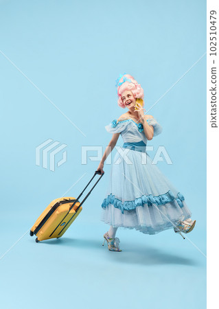 Portrait with happy young princess, queen wearing big pink wig holding suitcase talking via cell phone on blue background Portrait with happy young princess, queen wearing big pink wig holding suitcase talking via cell phone on blue background 102510479