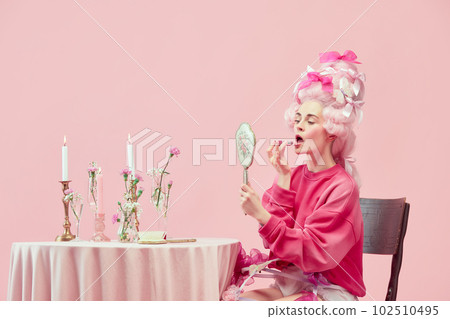 Portrait with pretty princess, queen wearing big wig holding hand mirror and using lipstick on pink background. Beauty salon 102510495