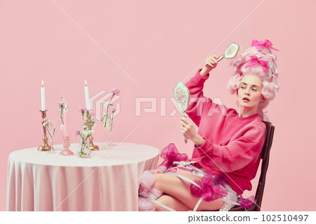 Portrait with princess wearing fancy pink clothes holding hand mirror and doing hairstyle over pink background. Beauty salon 102510497